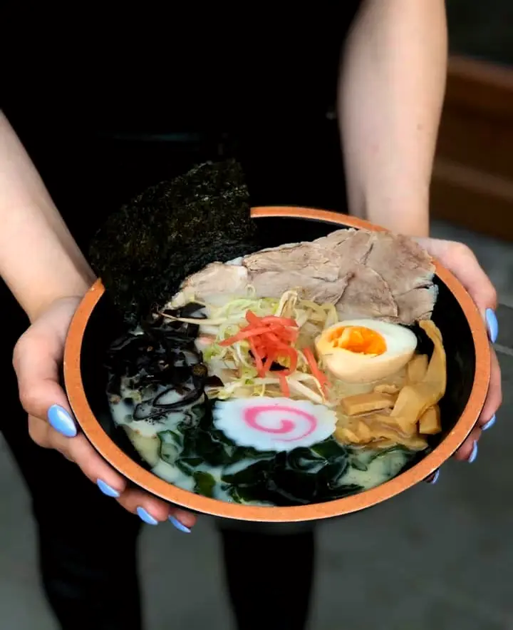 Dłonie podają miskę ramenu z narutomaki, jajkiem i chashu